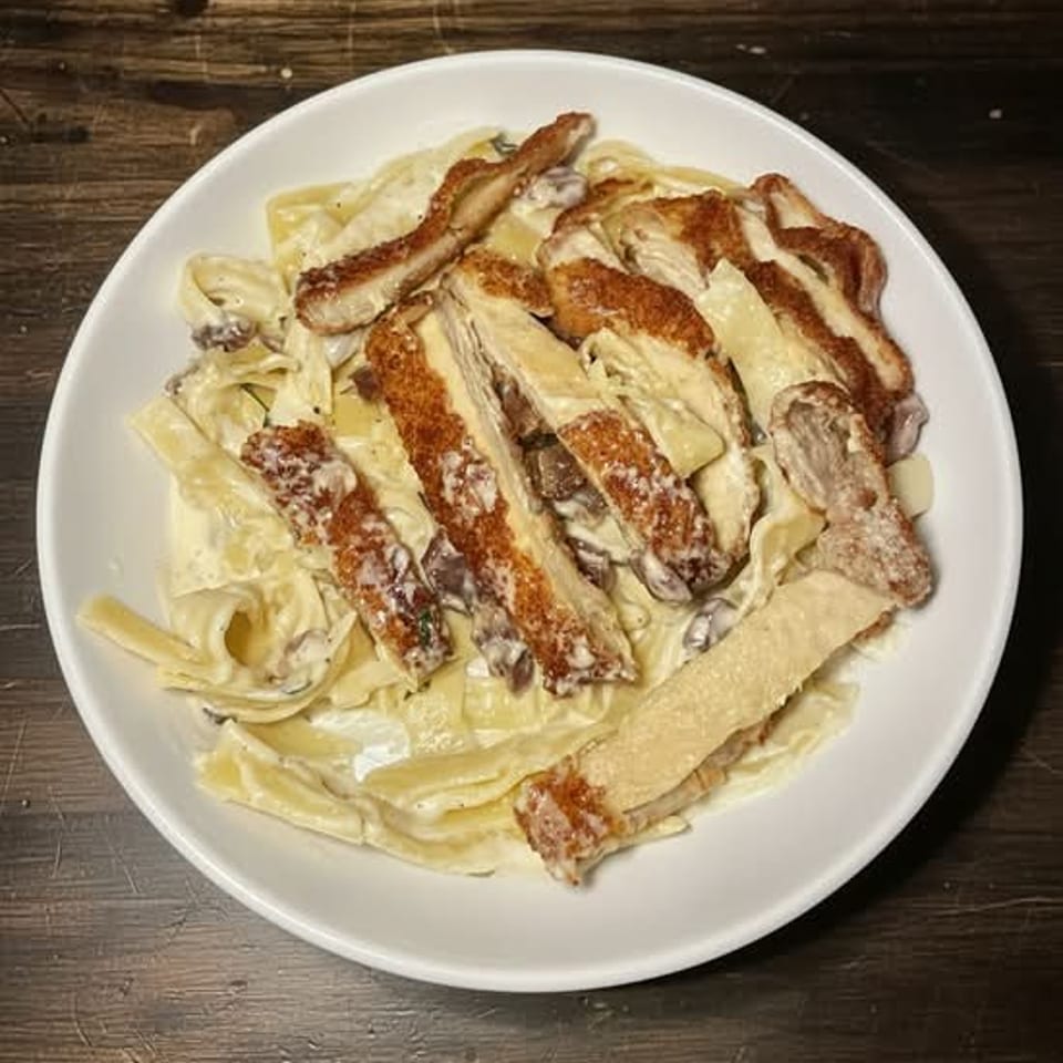 Creamy pasta with mushrooms and crispy chicken strips in a white bowl on a wooden table.