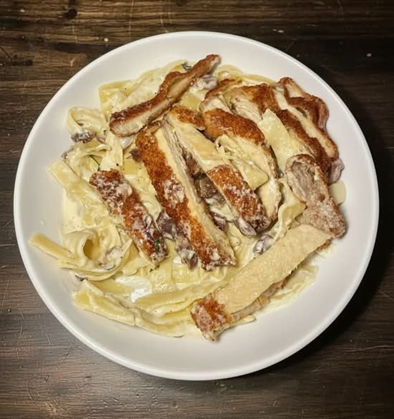 Creamy pasta with mushrooms and crispy chicken strips in a white bowl on a wooden table.