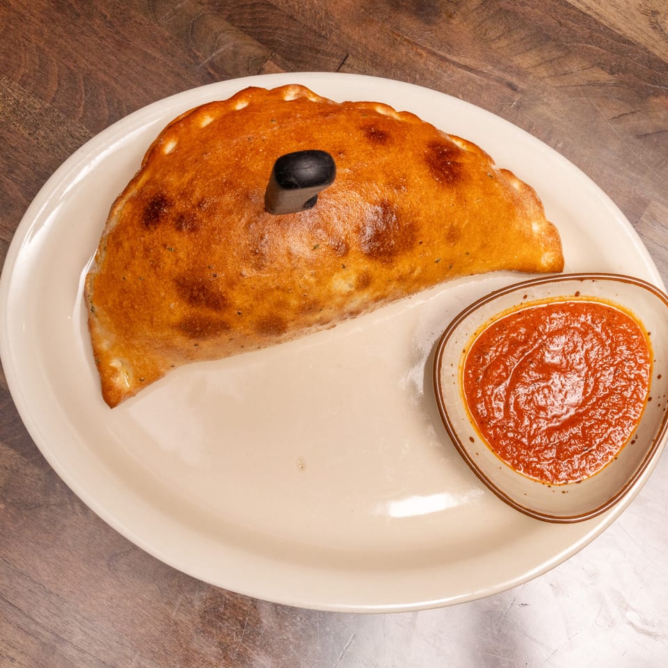 Golden-brown calzone with a knife, served on a white plate with a side of red marinara sauce.