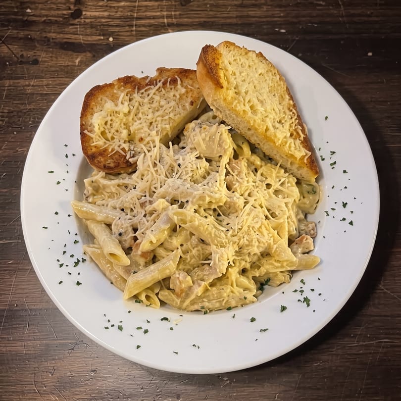 Creamy penne pasta with shredded cheese and parsley, served with two slices of cheesy garlic bread.