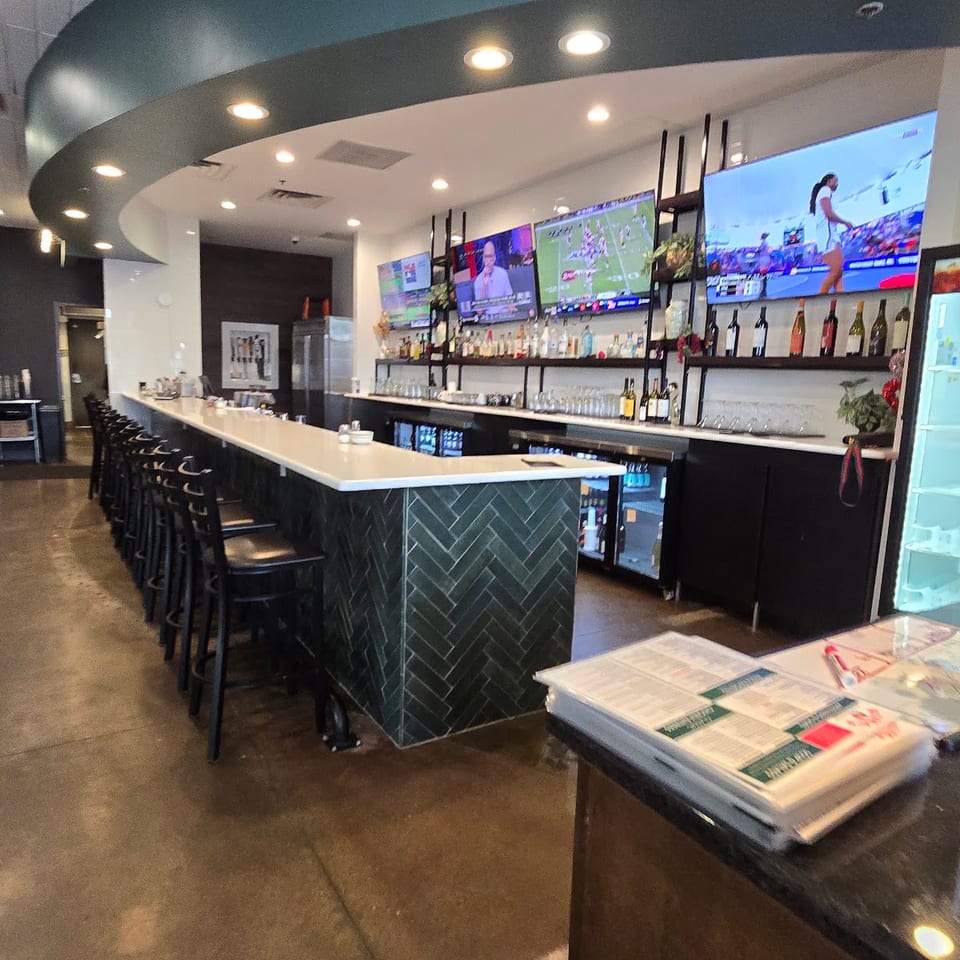 A long, empty bar with black stools, multiple TVs, and bottles of liquor on shelves.
