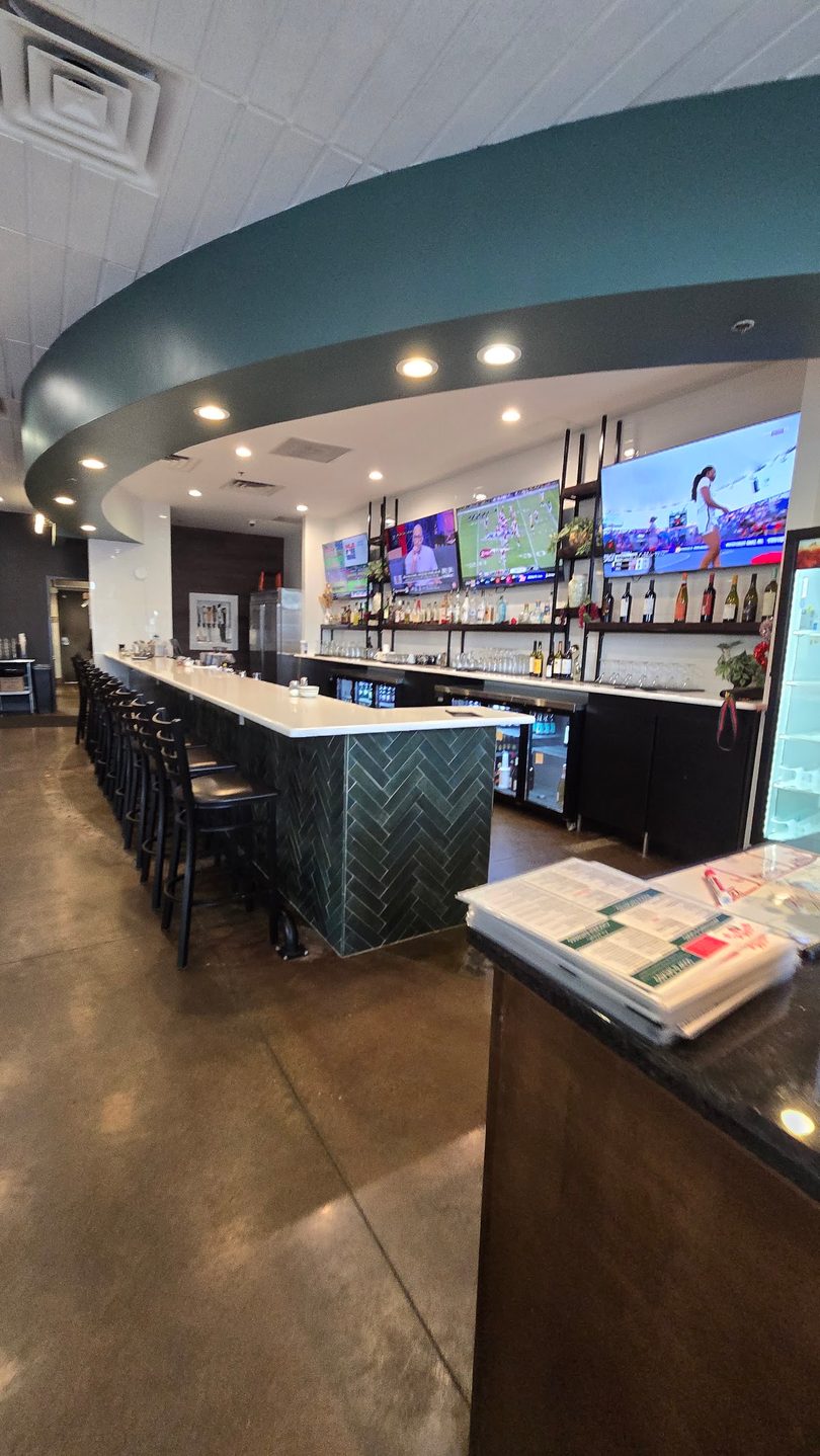 A long, empty bar with black stools, multiple TVs, and bottles of liquor on shelves.