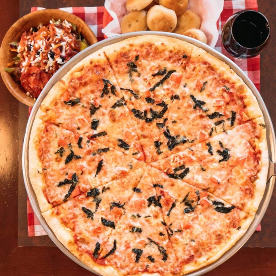 A large Margherita pizza with basil, salad, bread rolls, and red wine on a checkered tablecloth.