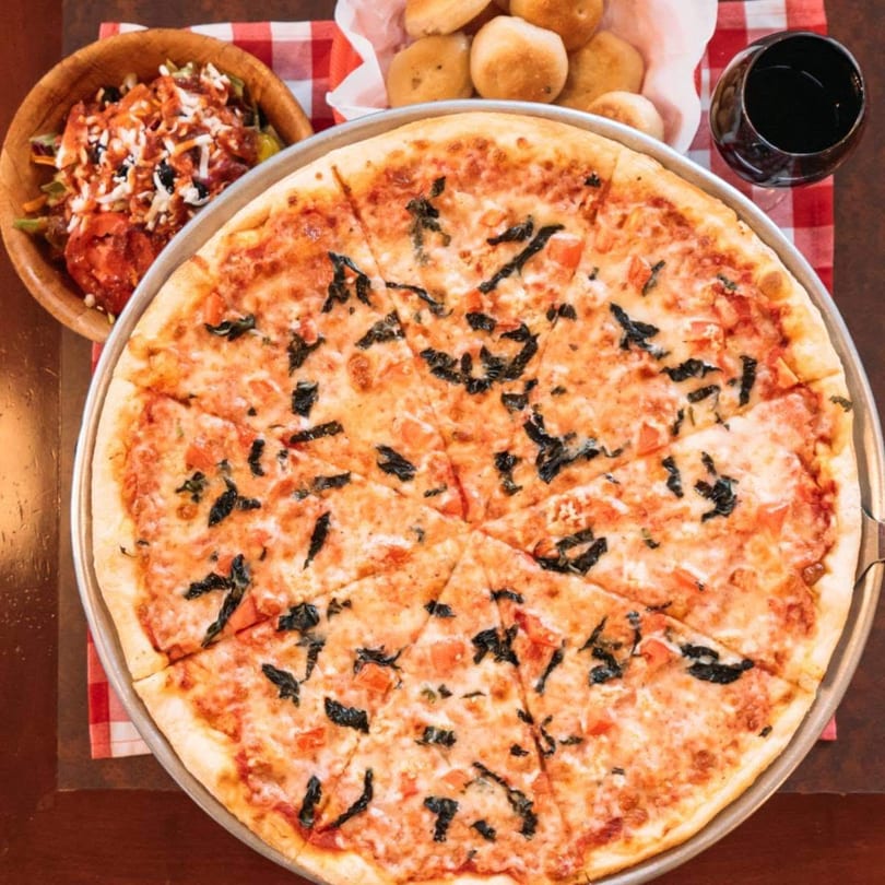 A large Margherita pizza with basil, salad, bread rolls, and red wine on a checkered tablecloth.