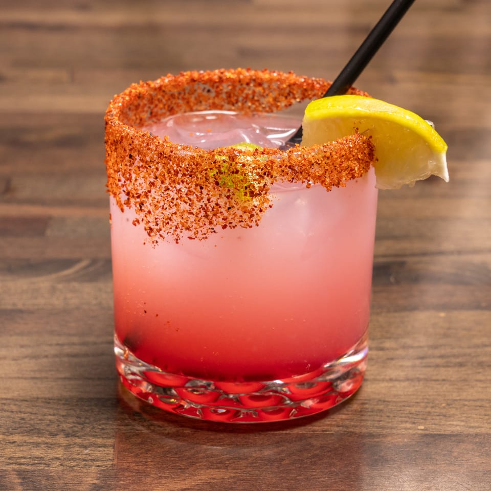 Pink cocktail with a chili-lime rim, ice, a lemon wedge, and a black straw on a wooden table.
