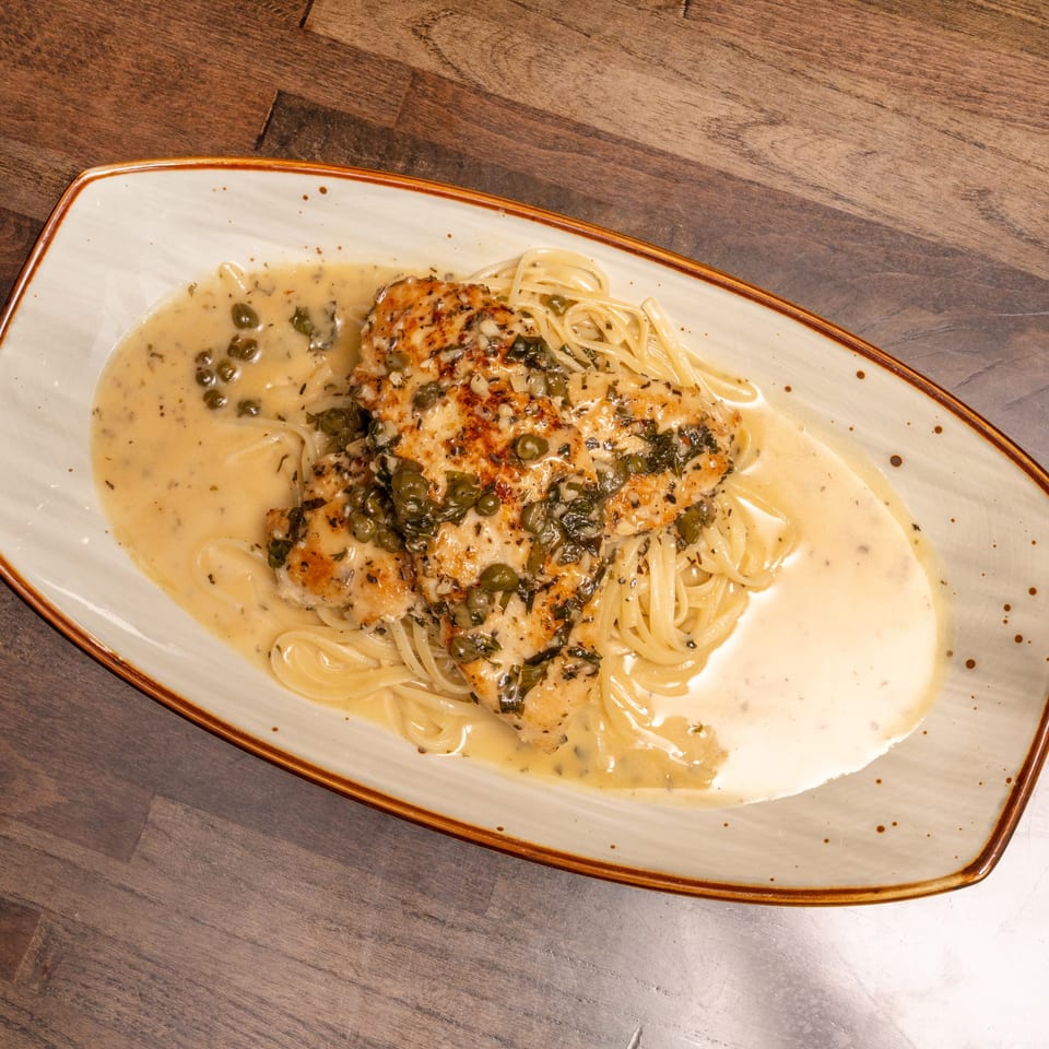 Chicken piccata with capers and herbs over spaghetti in a creamy sauce, on a rectangular plate.