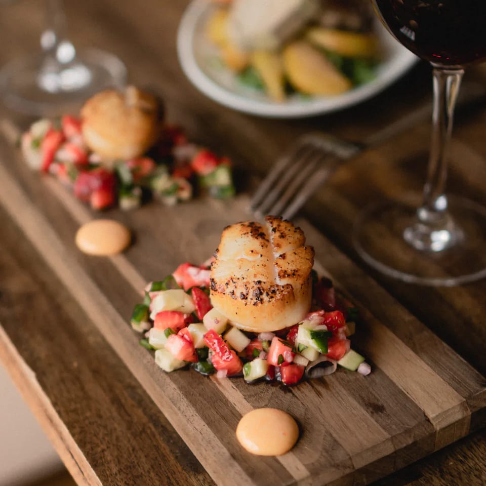 Seared scallops with strawberry and cucumber salsa, served on a wooden board with wine glasses.