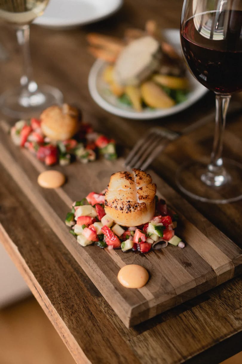Seared scallops with strawberry and cucumber salsa, served on a wooden board with wine glasses.