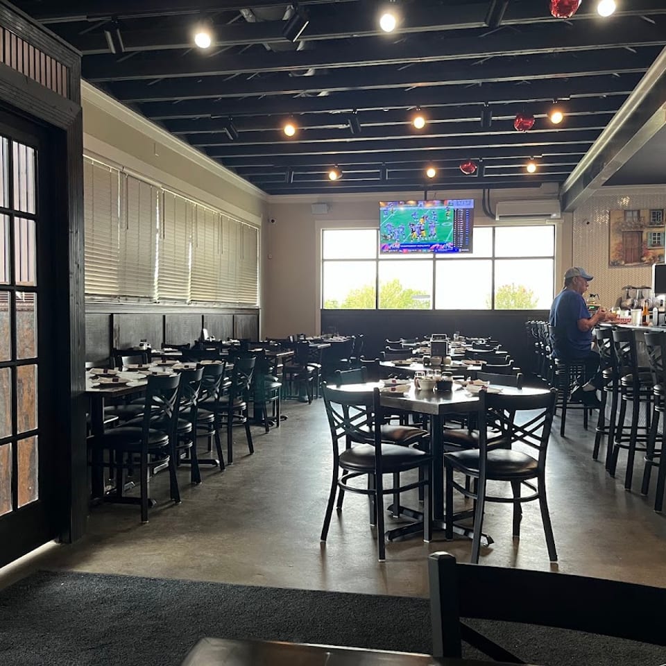 Spacious restaurant interior with dark tables, chairs, a bar, and a man watching TV.