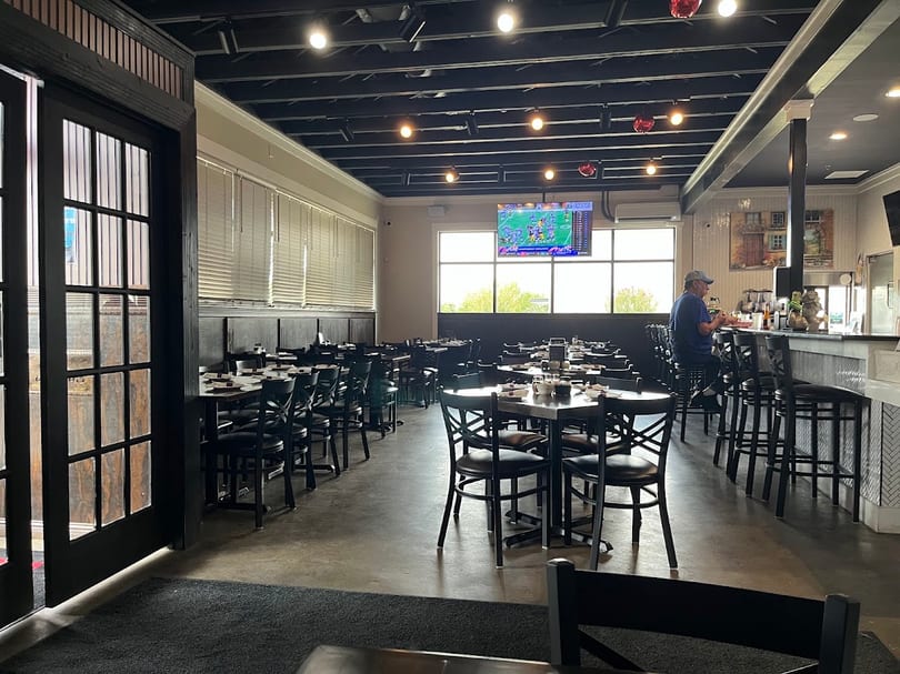 Spacious restaurant interior with dark tables, chairs, a bar, and a man watching TV.