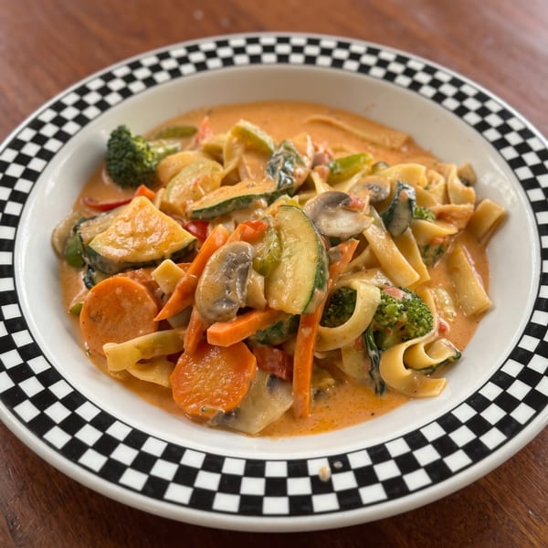 Pasta with creamy sauce, zucchini, carrots, mushrooms, and broccoli in a checkered bowl.