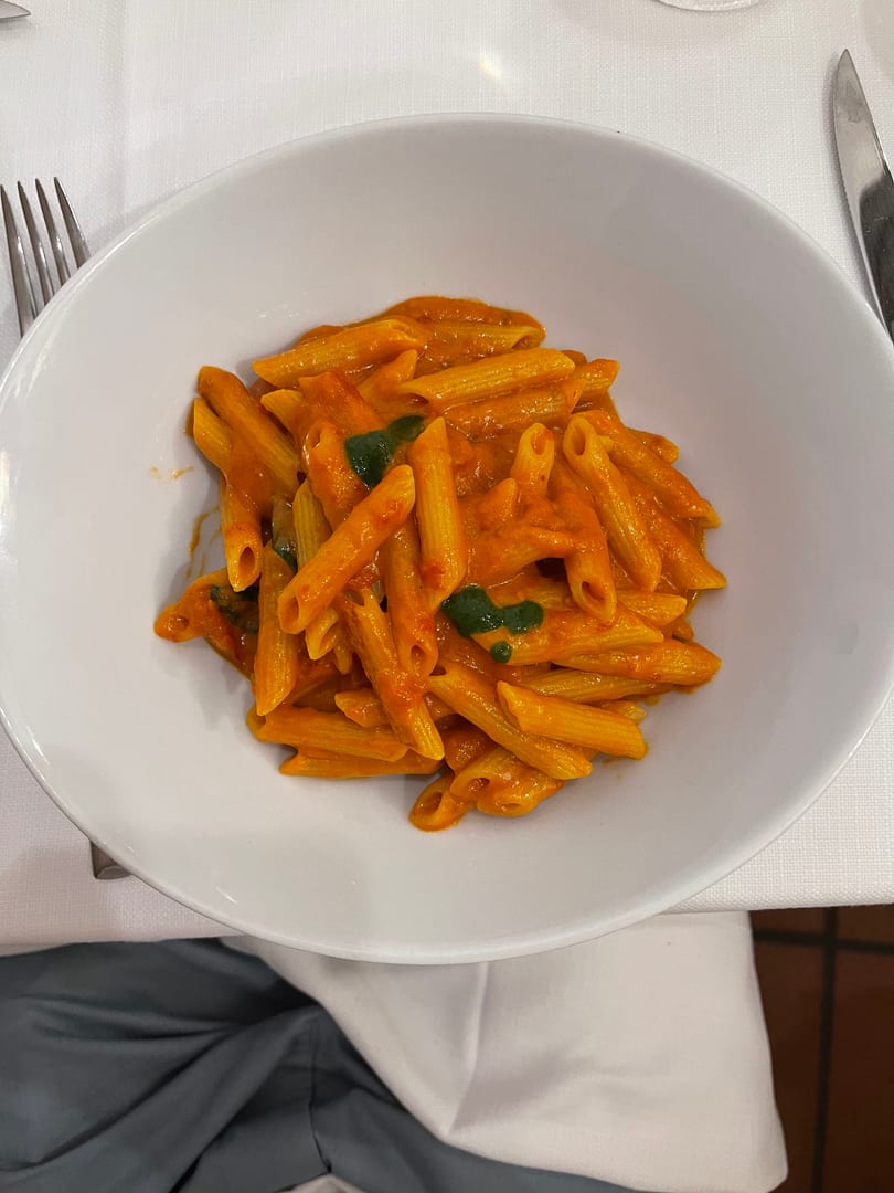 Penne pasta in a creamy orange sauce with green herbs in a white bowl on a white tablecloth.