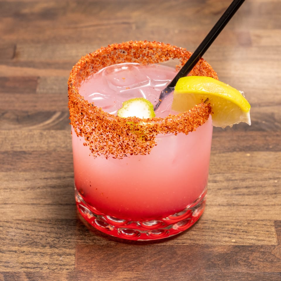 Pink cocktail with a chili-lime rim, lemon and lime wedges, and a black straw on a wooden table.