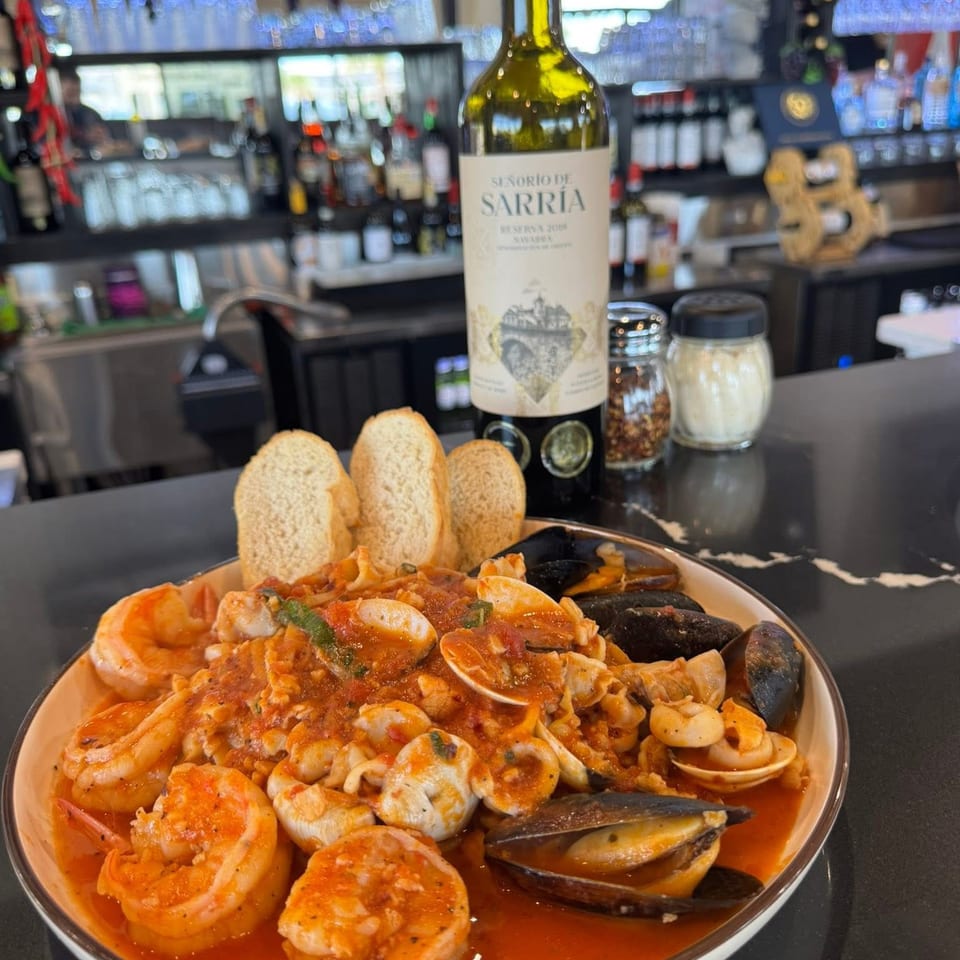 Seafood pasta with shrimp, mussels, and clams in red sauce, bread, and wine bottle on a bar.