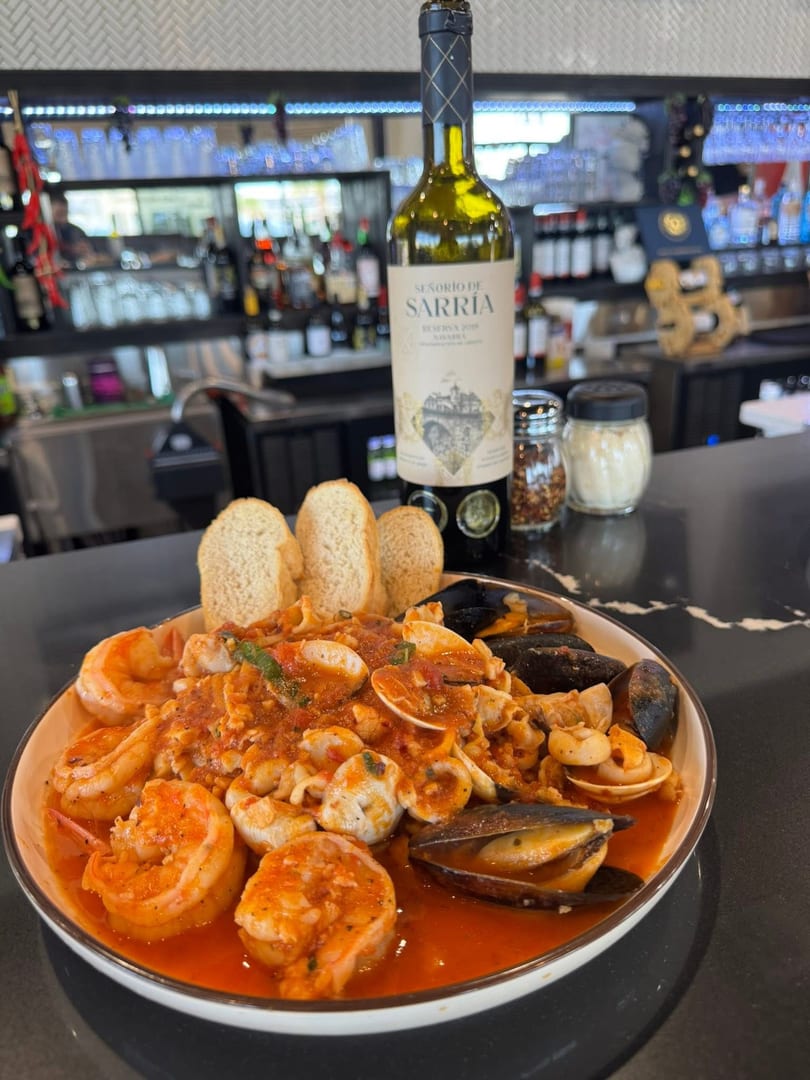 Seafood pasta with shrimp, mussels, and clams in red sauce, bread, and wine bottle on a bar.