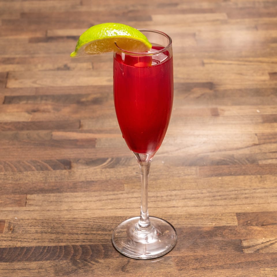A vibrant red cocktail in a champagne flute, garnished with a fresh lime wedge on a wooden table.