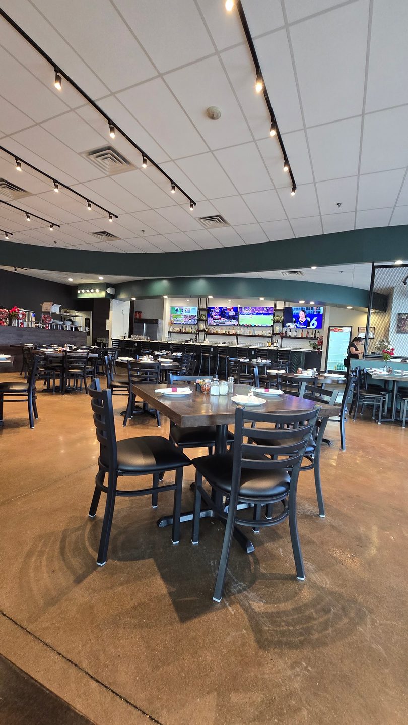 A restaurant dining room with dark tables and chairs, a bar with TVs, and a person standing near the bar.