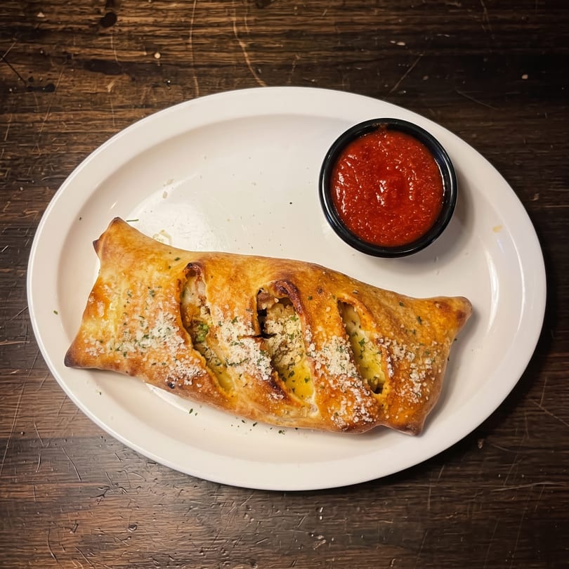 Golden-brown calzone topped with herbs and cheese, served with a side of red sauce on a white plate.