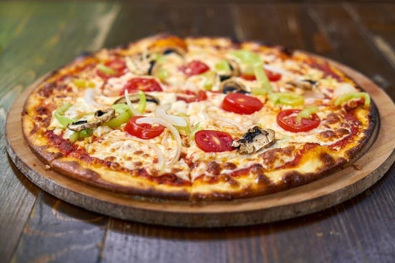 Freshly baked pizza with melted cheese, tomatoes, green peppers, onions, and mushrooms on a wooden board.
